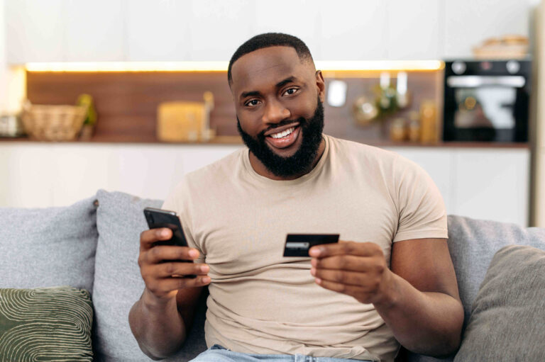 Happy african american attractive man, sits on a sofa in the living room, holding a bank card and a smartphone, making online purchases, making payments, ordering delivery
